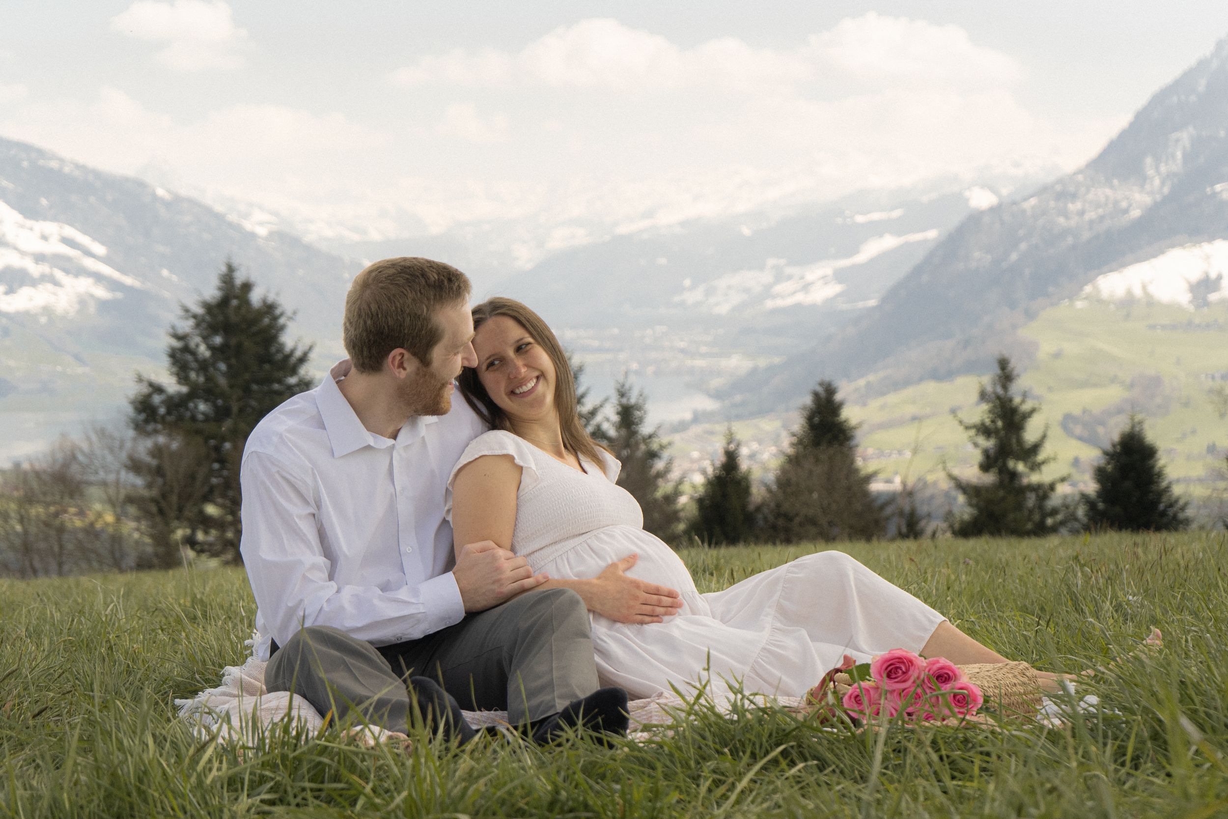 Pregnant couple sitting on a grassy alpine meadow with snowy mountains in the background on a cloudy day.