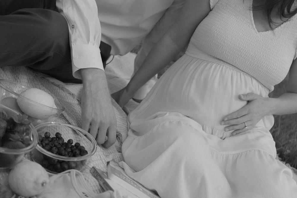 Black and white photo of a pregnant woman in white dress with partner at a picnic, hands on baby bump.