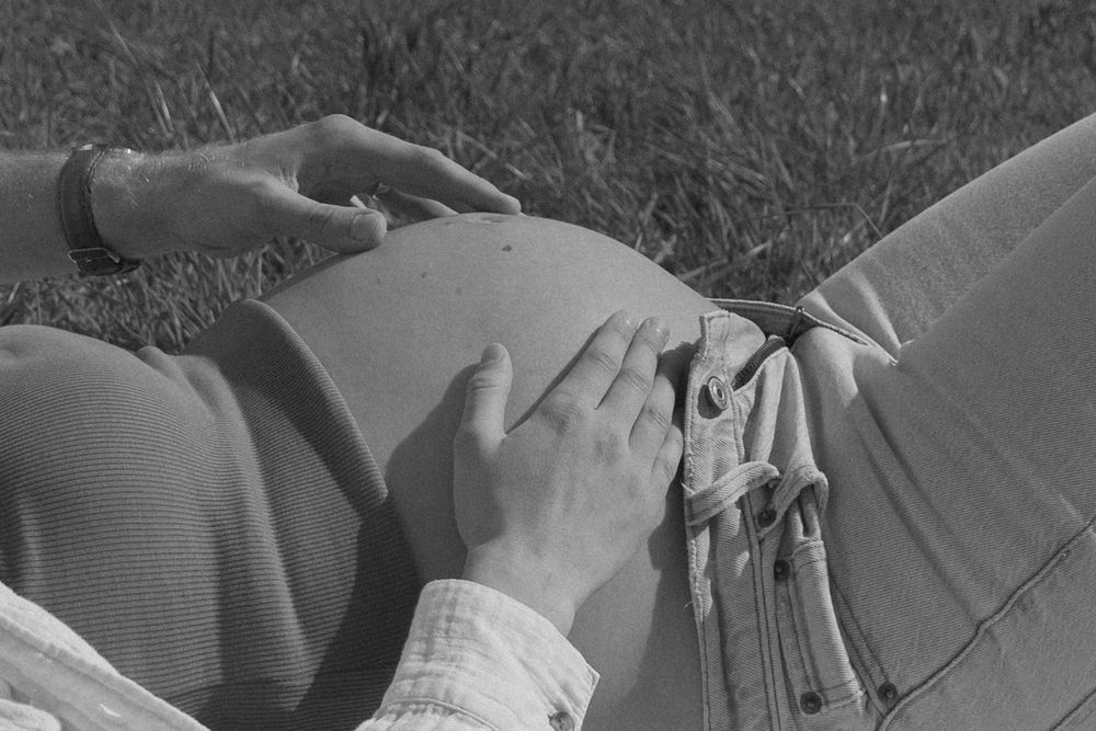 Black and white photo of a pregnant woman lying on grass, gently cradling her baby bump with both hands.