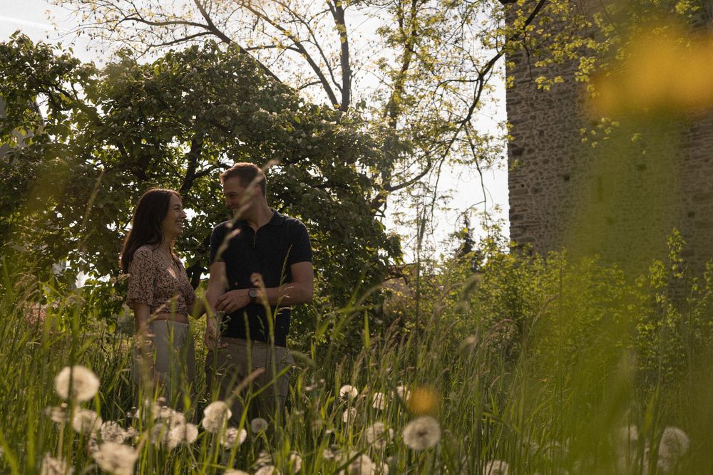 Couple standing together in a lush green meadow with wildflowers and tall trees in warm golden sunlight.