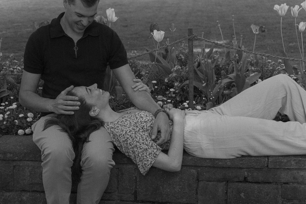 Black and white photo of a couple relaxing in a flower garden, woman lying down smiling up at man seated beside her.
