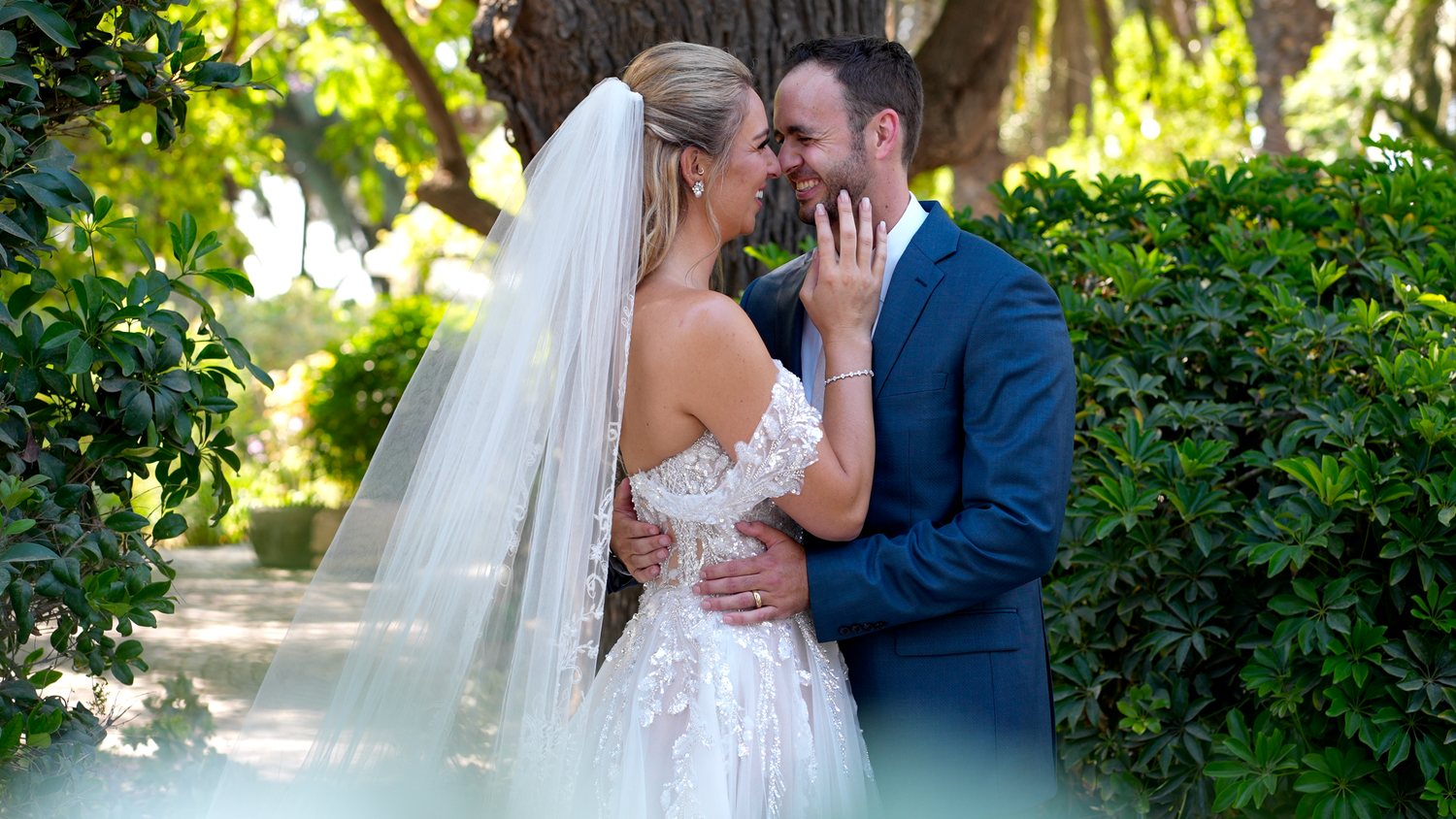 A romantic wedding moment captured in a lush garden setting with sunlight filtering through green foliage.