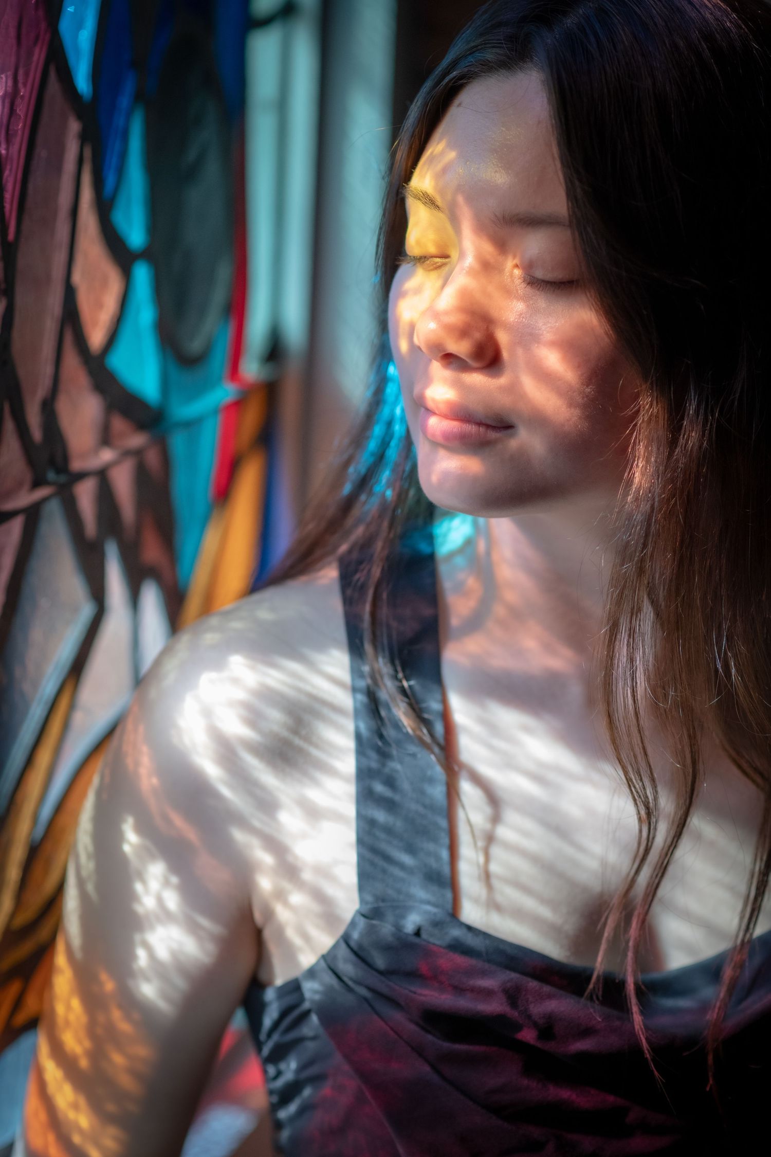 Young woman with eyes closed, colorful light patterns on her face near stained glass.