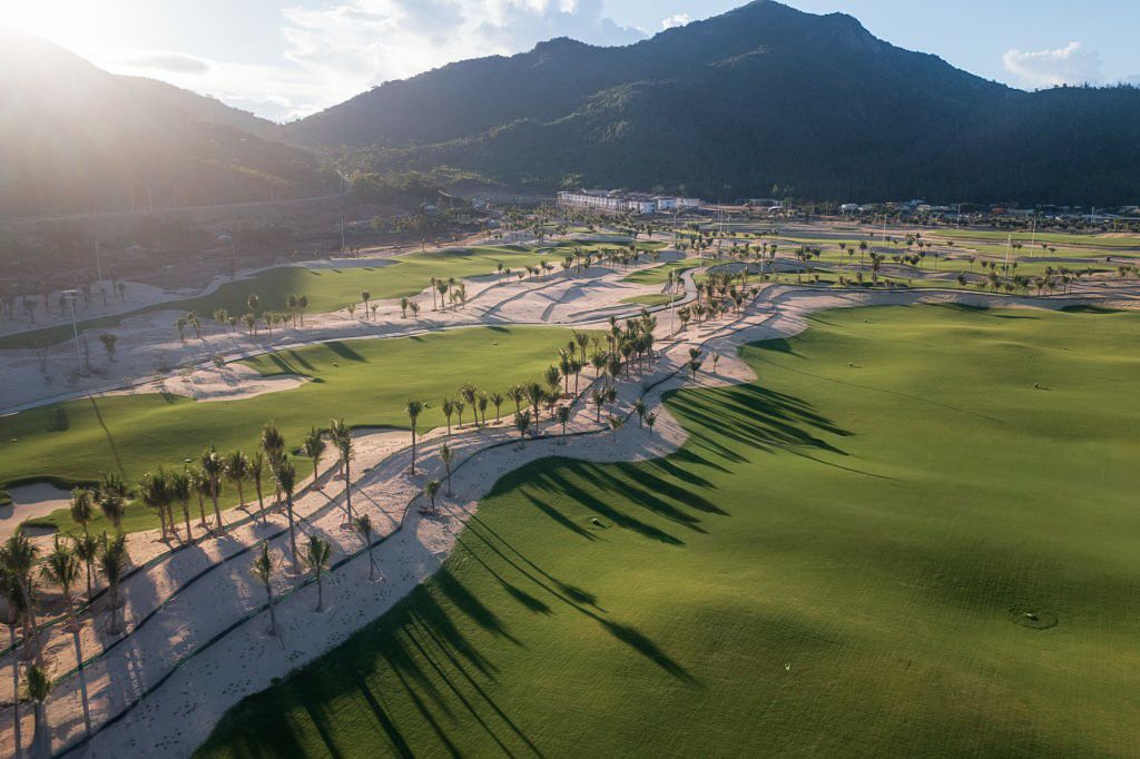 Aerial panoramic view of a golf course nestled in a valley surrounded by majestic mountain peaks at sunset.