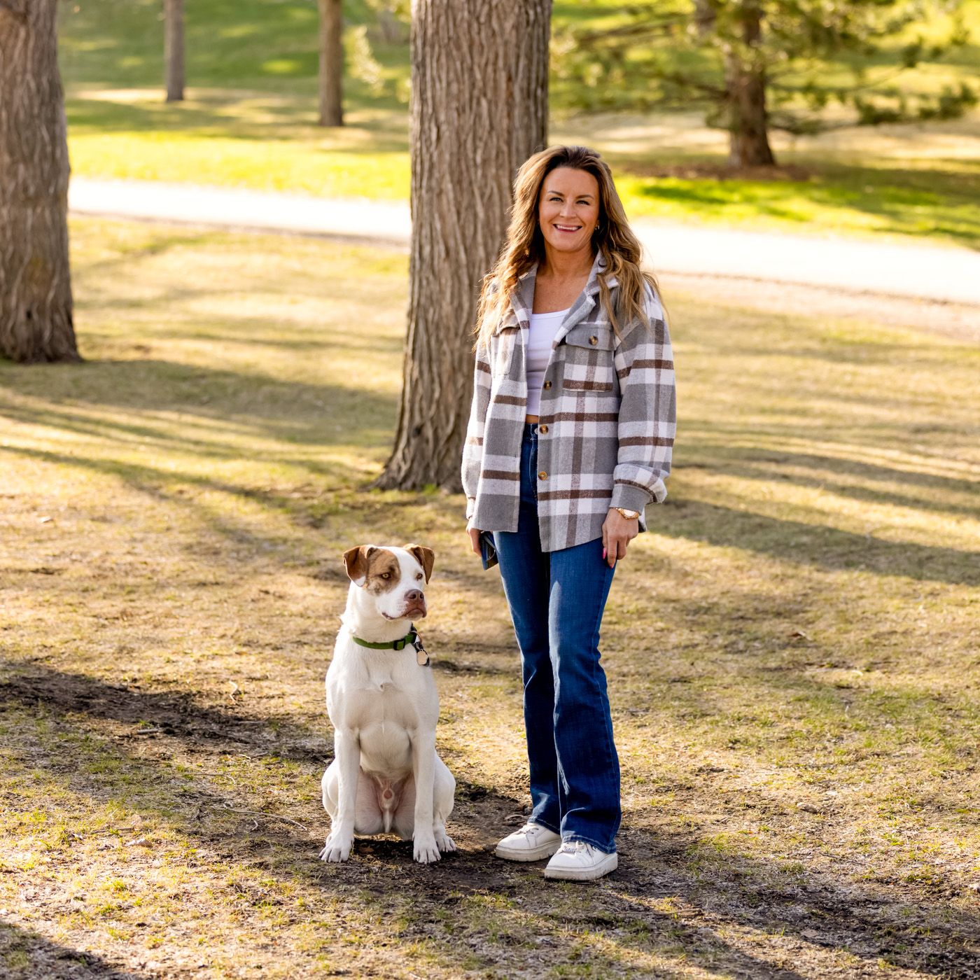 A white dog sits obediently in a sunny park setting while being trained with a plaid-shirted companion.