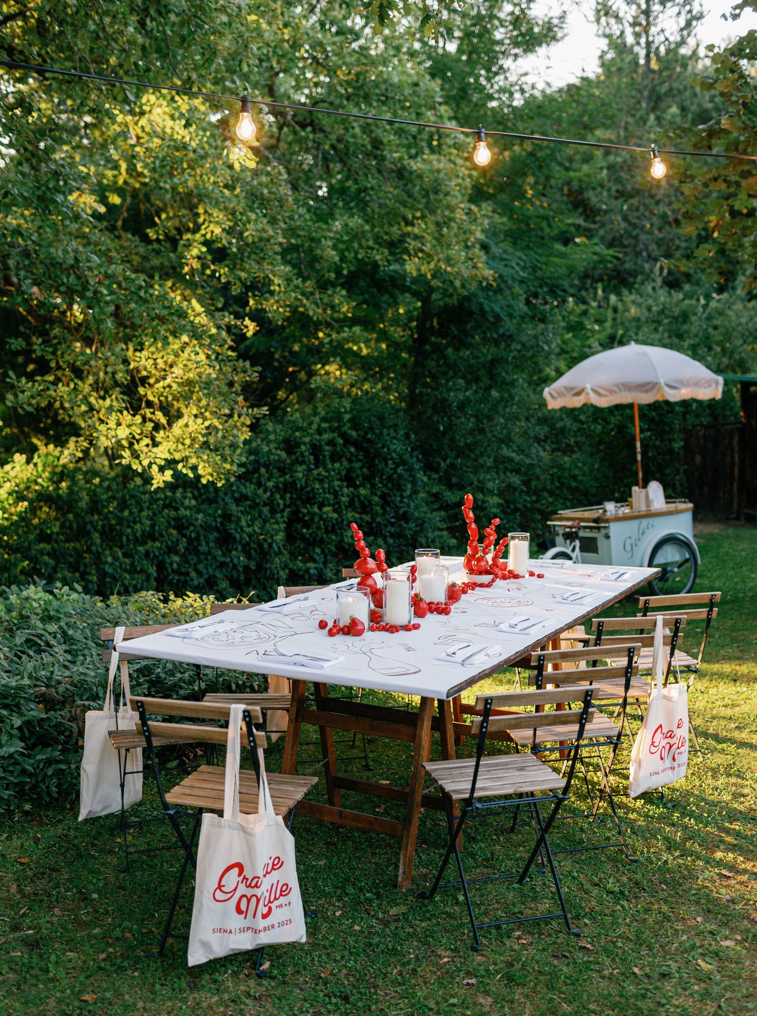 Long banquet tables decorated with white linens and red accents set up for outdoor dining under string lights.