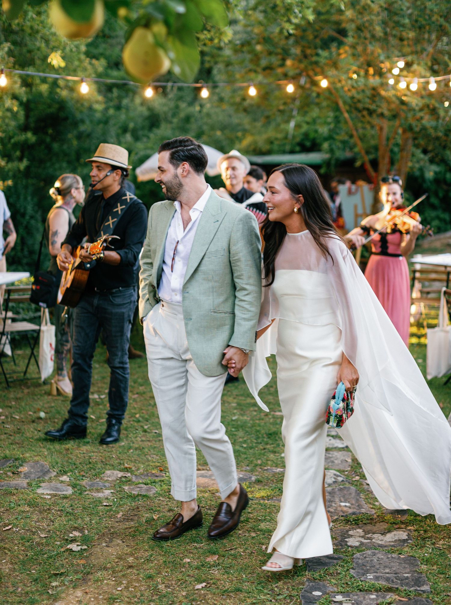 Wedding guests dance and celebrate under string lights at an outdoor garden reception in the evening.