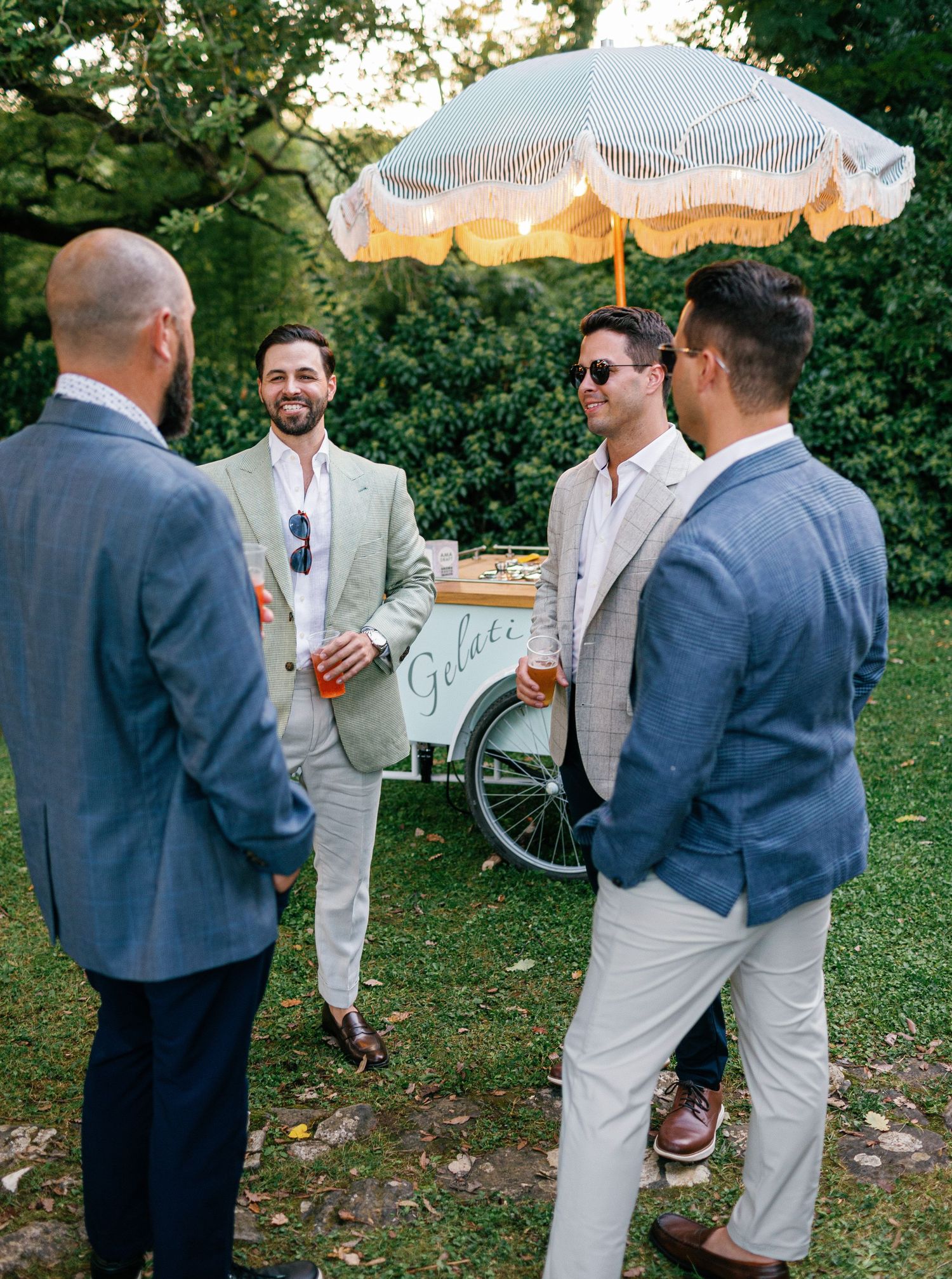 Group of people chatting and socializing outdoors at a garden party wearing light colored and navy suits.