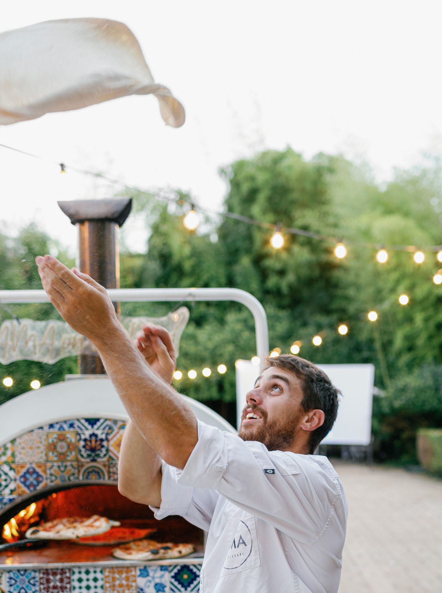 Three action shots show a chef skillfully tossing pizza dough in the air at an outdoor restaurant with string lights.