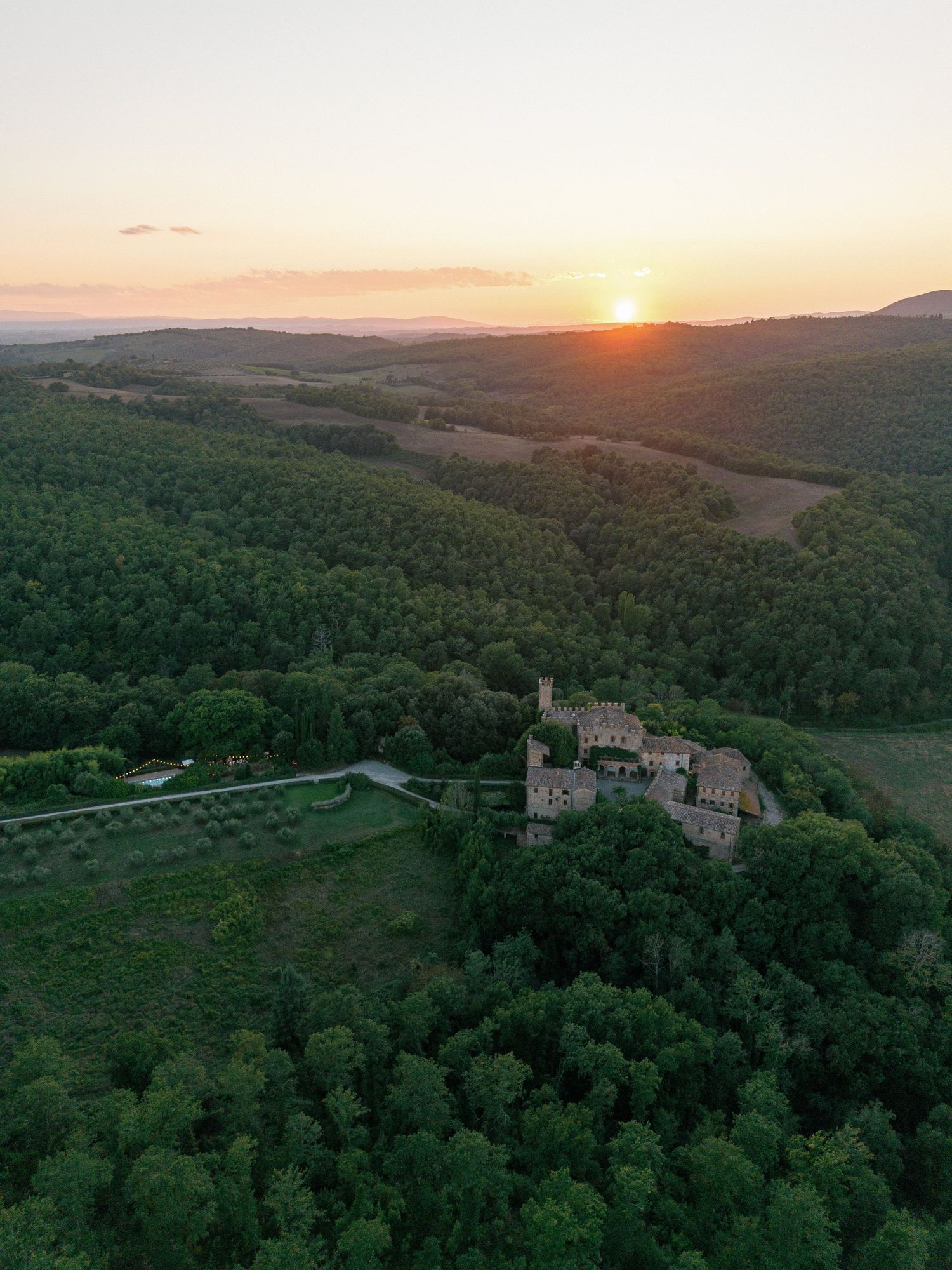 Aerial view of a medieval castle nestled in Tuscan hills during golden sunset over forested countryside.