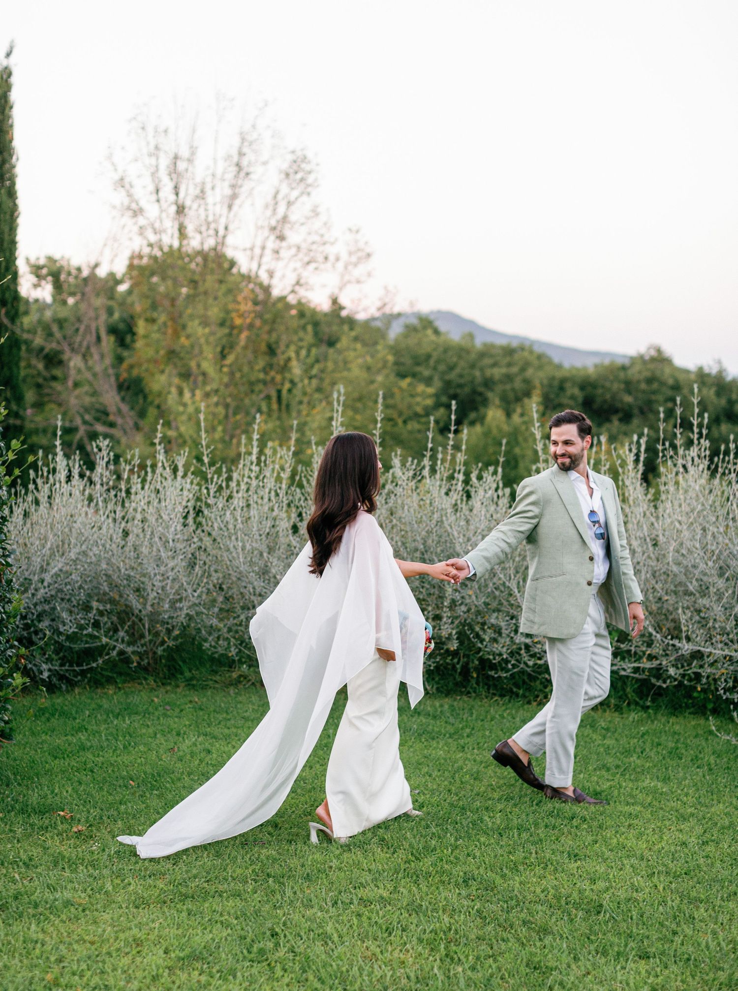 A couple in white outfits dances playfully through a garden with mountains and wildflowers in the background.