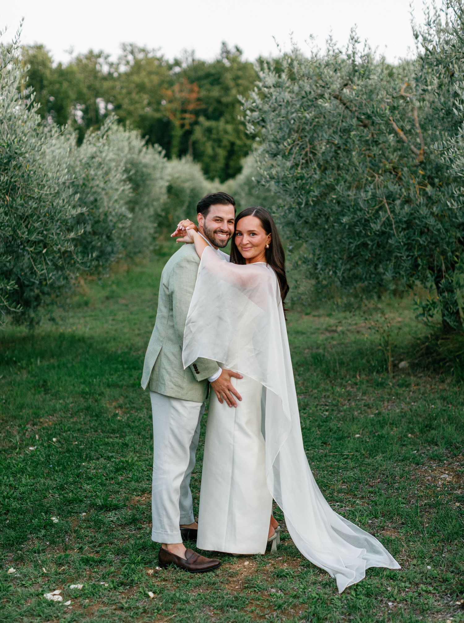 A couple in white outfits embraces while walking through a serene olive grove on a sunny afternoon.