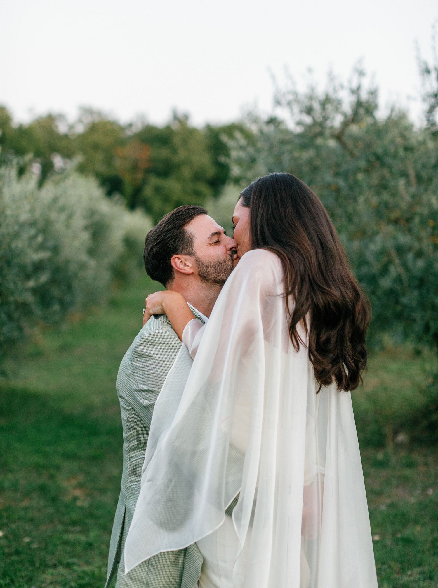 A romantic couple embraces in a dreamy outdoor setting with olive trees in the background.