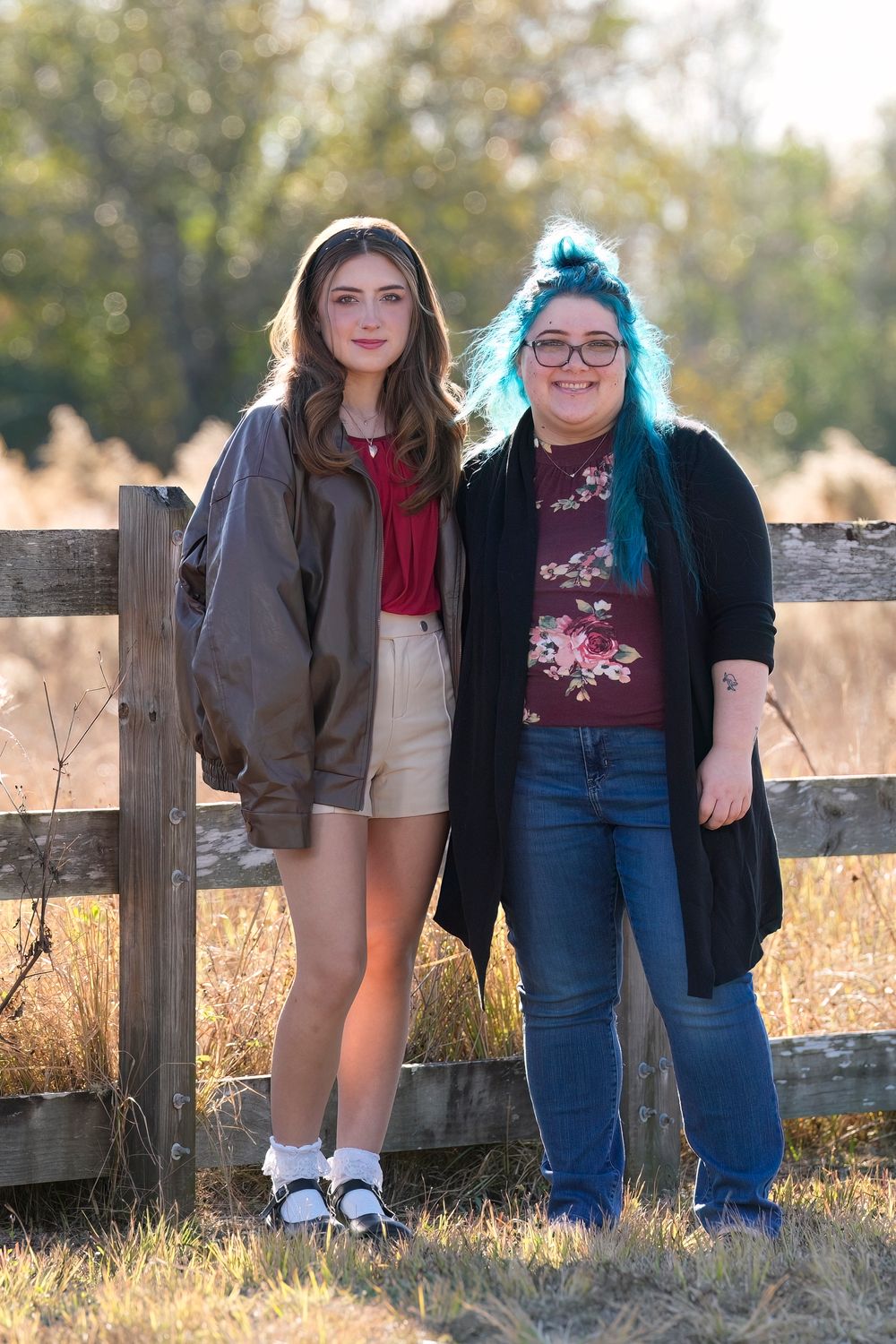 Two people stand together by a wooden fence in autumn sunlight wearing casual outfits and smiling.