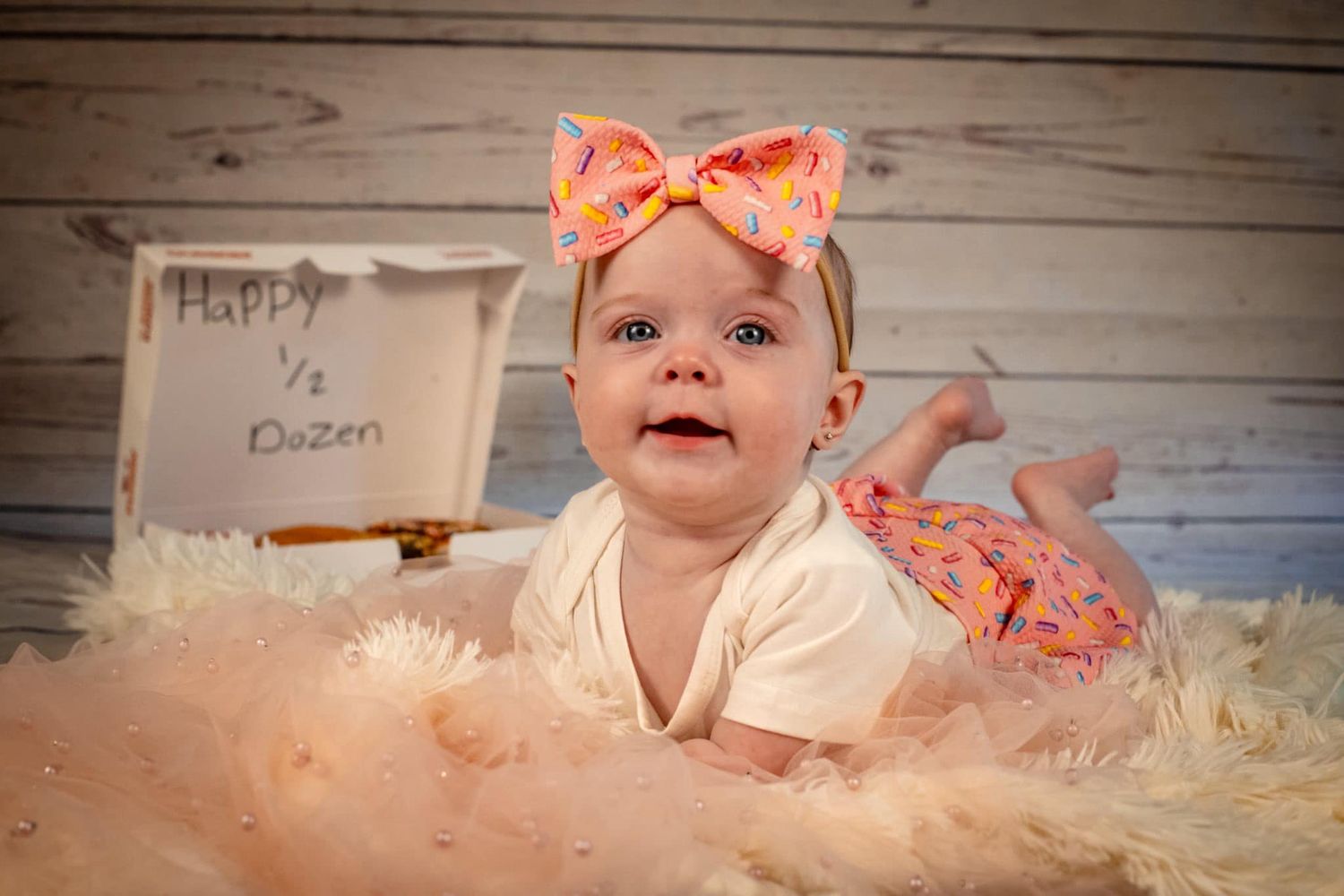 Baby wearing coral bow headband lying on fluffy rug during professional photoshoot.