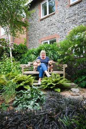 A stone cottage with a wooden bench surrounded by lush ferns and garden foliage creates a peaceful outdoor seating area.