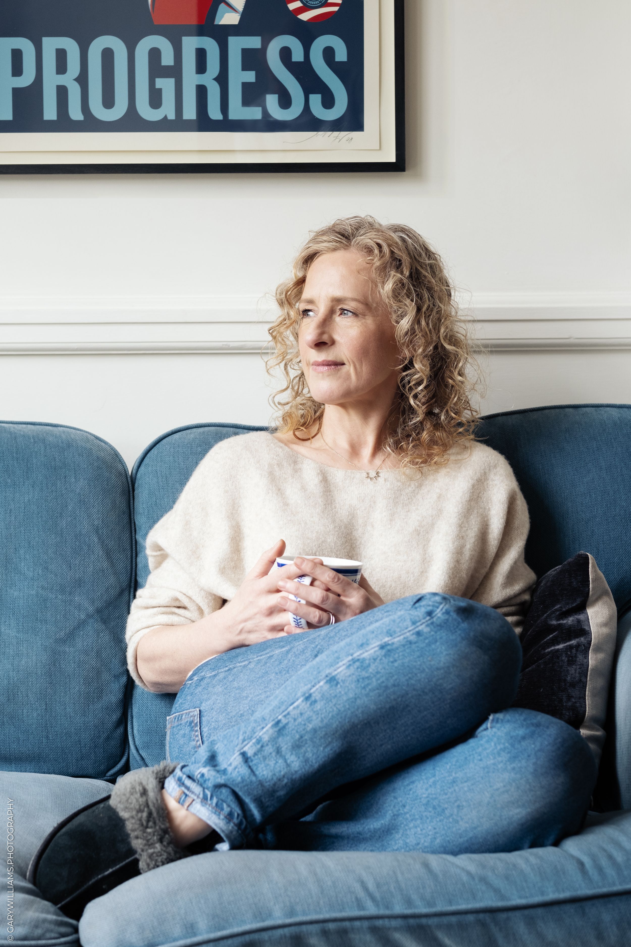 Person wearing white sweater and jeans relaxes on blue couch while looking out window in contemplative pose.