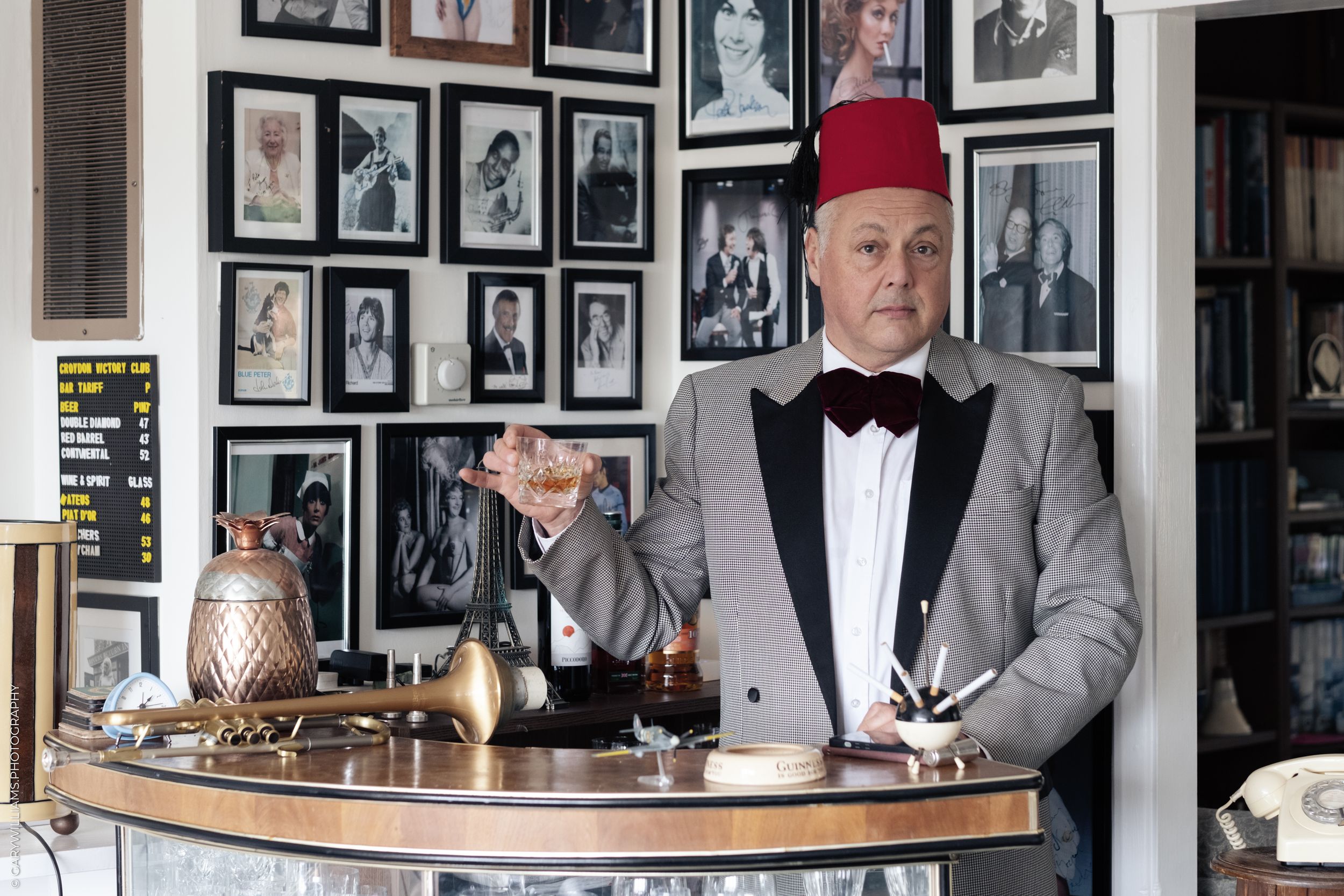 A bartender in a red fez hat and tuxedo stands behind a wooden bar counter with vintage photos on the wall behind him.