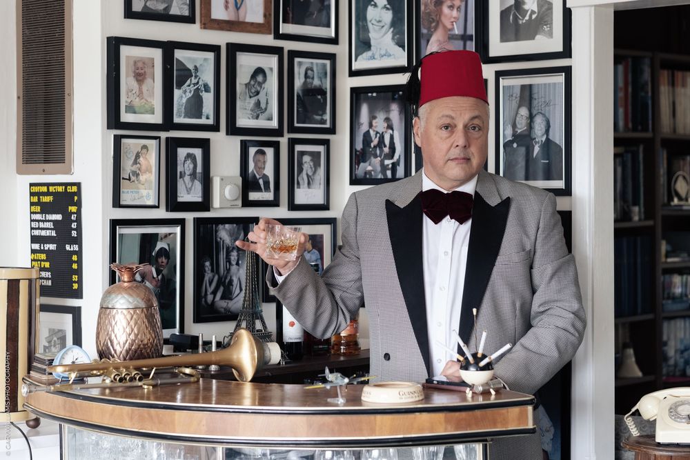 A bartender in a red fez hat and tuxedo stands behind a wooden bar counter with vintage photos on the wall behind him.