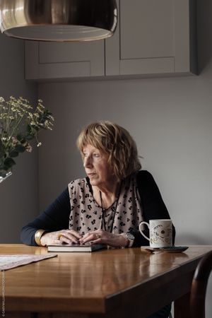 Person sits at wooden table with coffee mug and flowers, gazing thoughtfully by window in moody lighting.