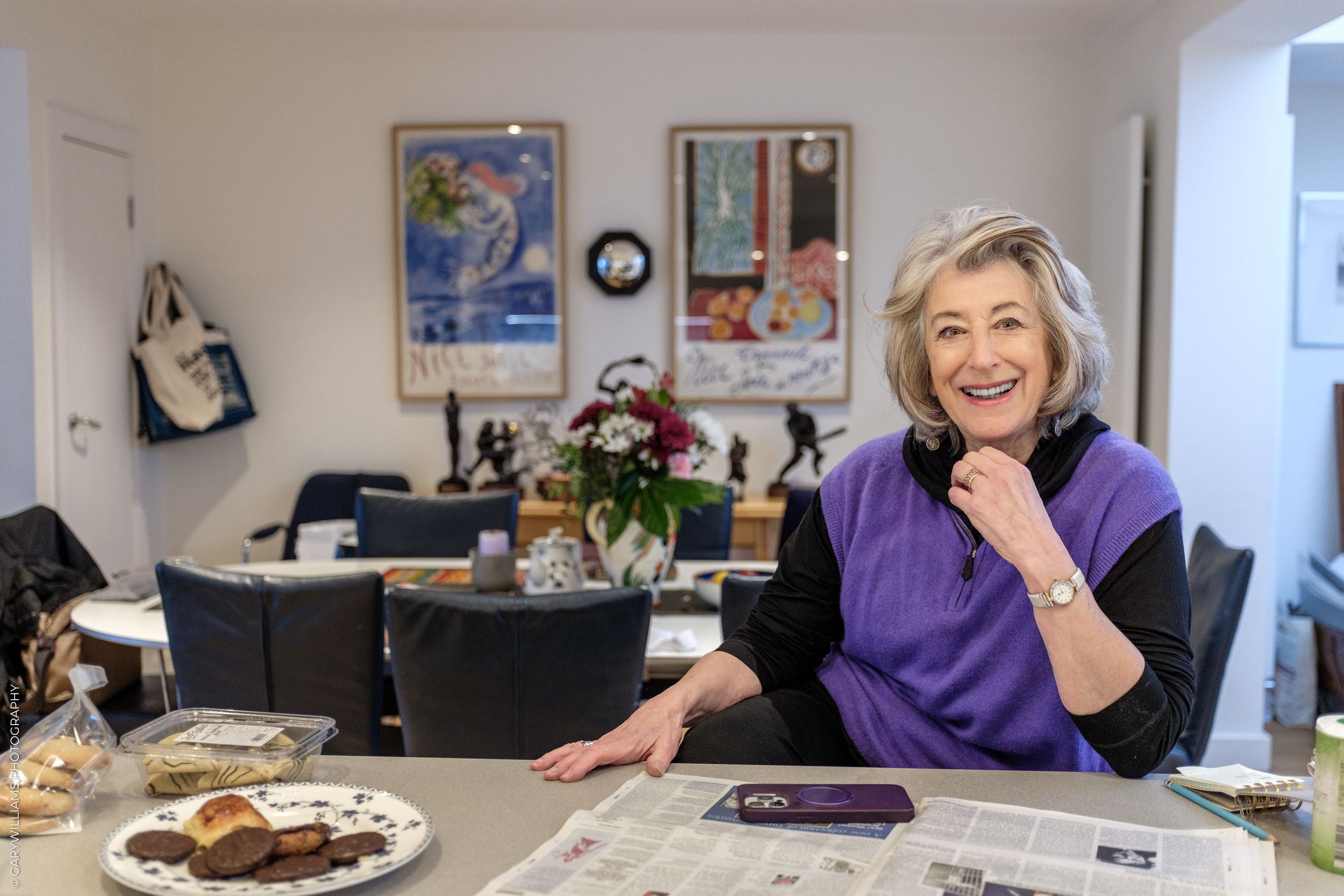 Maureen Lipman in a purple sweater sits at a dining table with cookies and flowers, laughing in a bright room with art on the walls.