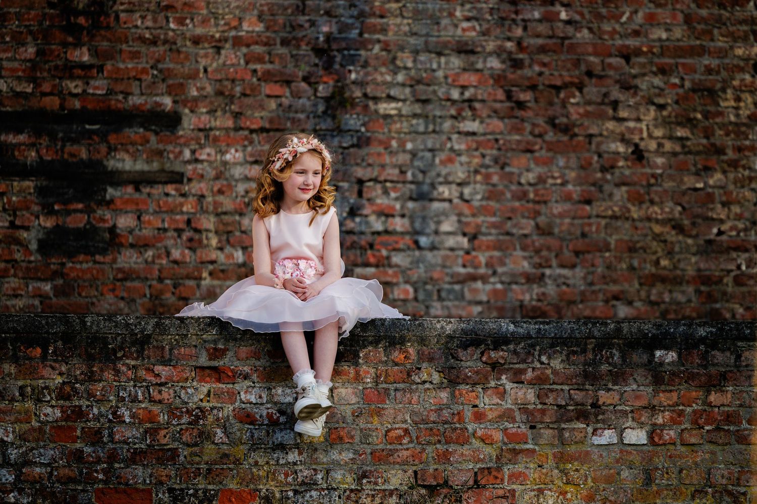 Ballet dancer in pink tutu sits gracefully against rustic brick wall during outdoor portrait session.