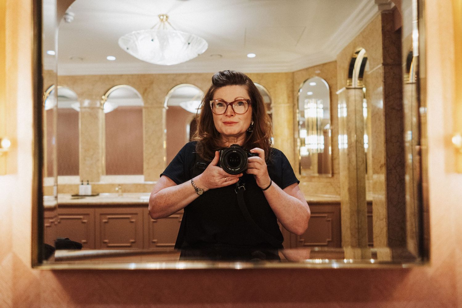A person wearing glasses stands in an elegant hallway with ornate mirrors and chandeliers.