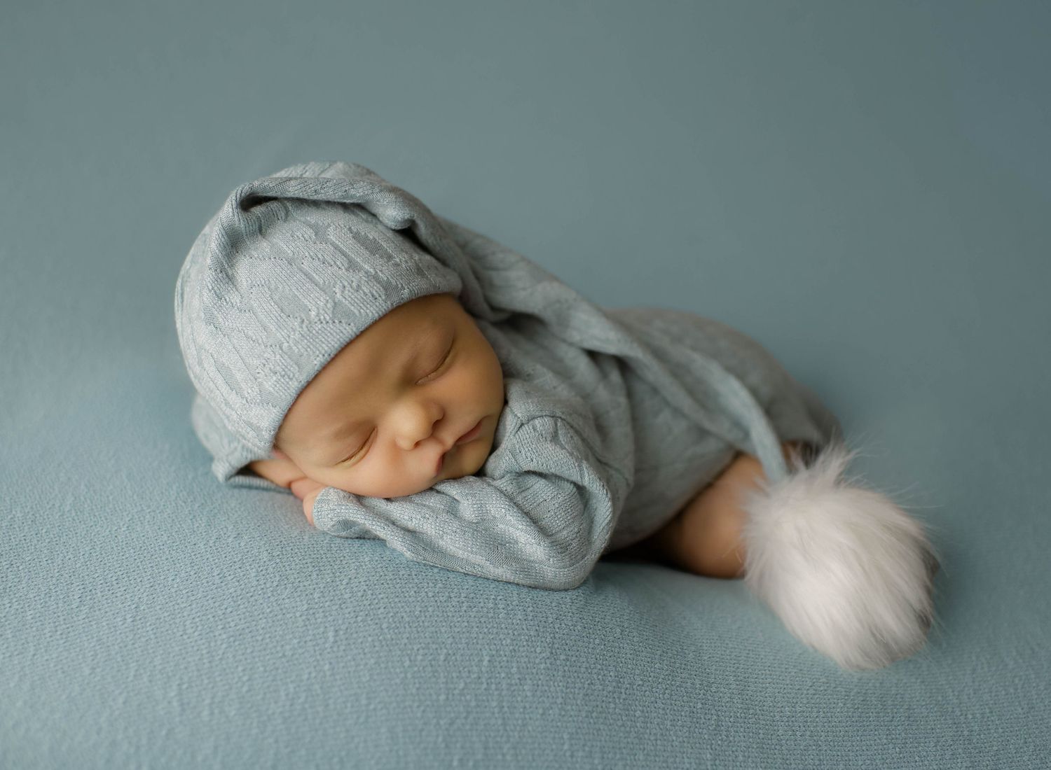 Sleeping newborn wearing soft gray knit outfit with pom pom hat on blue backdrop.