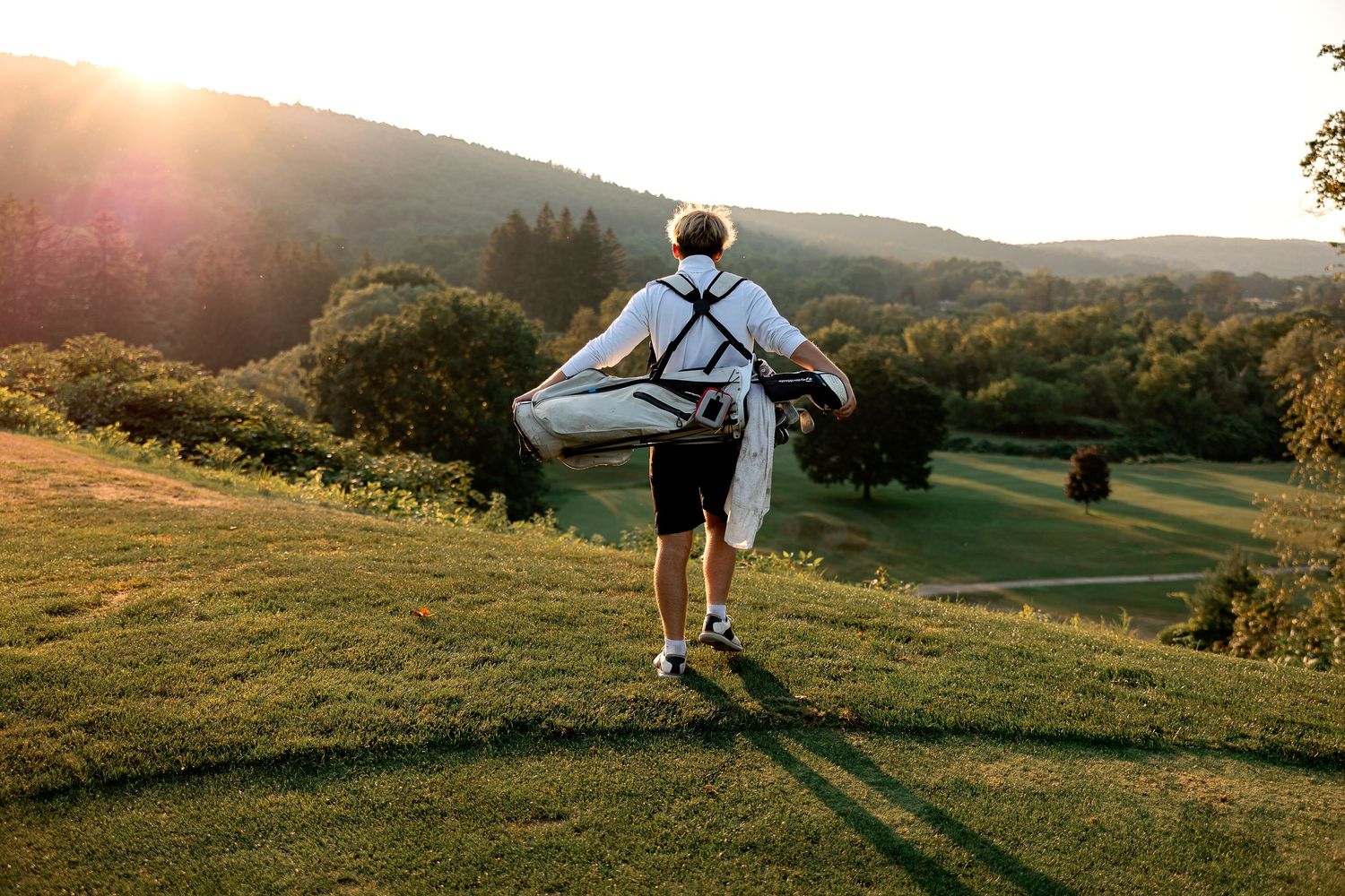 A hiker with a backpack walks across a grassy hillside at sunset with mountains in the background.