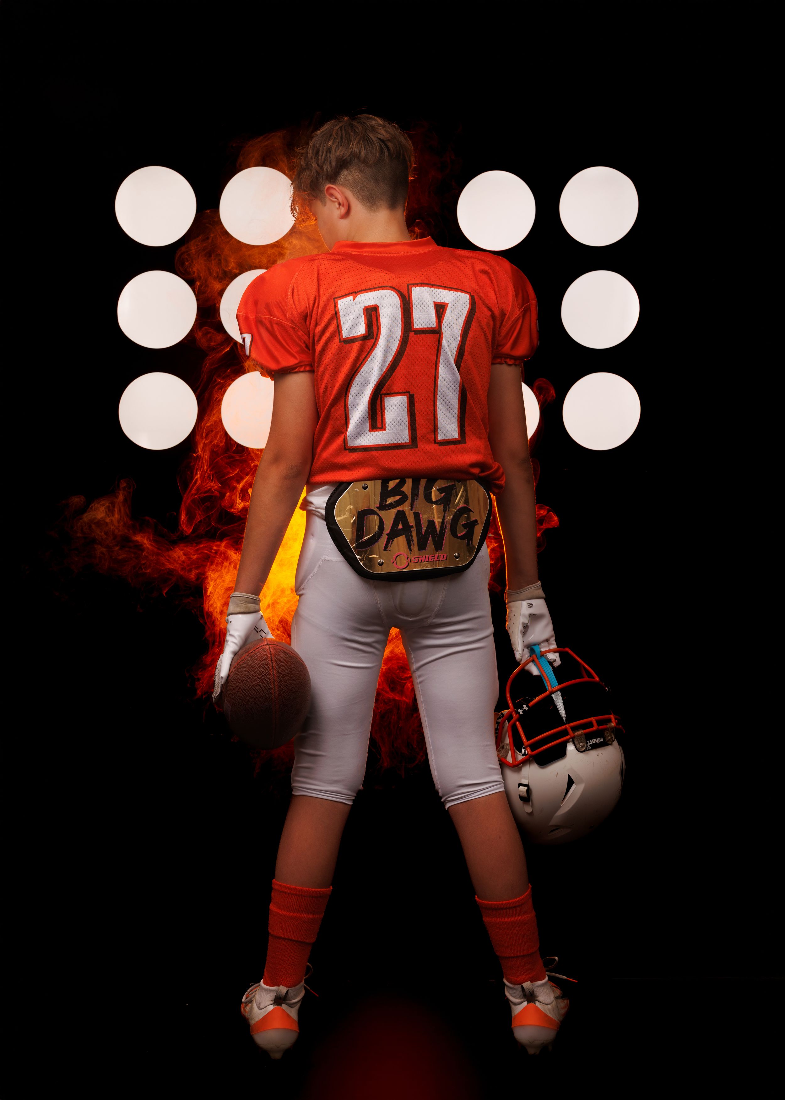 Softball player in orange jersey and gray pants photographed against dramatic black background with spotlights.