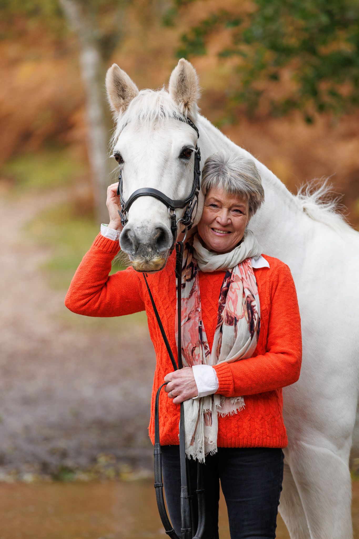 A beautiful white horse and someone in a bright orange sweater share an affectionate moment by a stream.