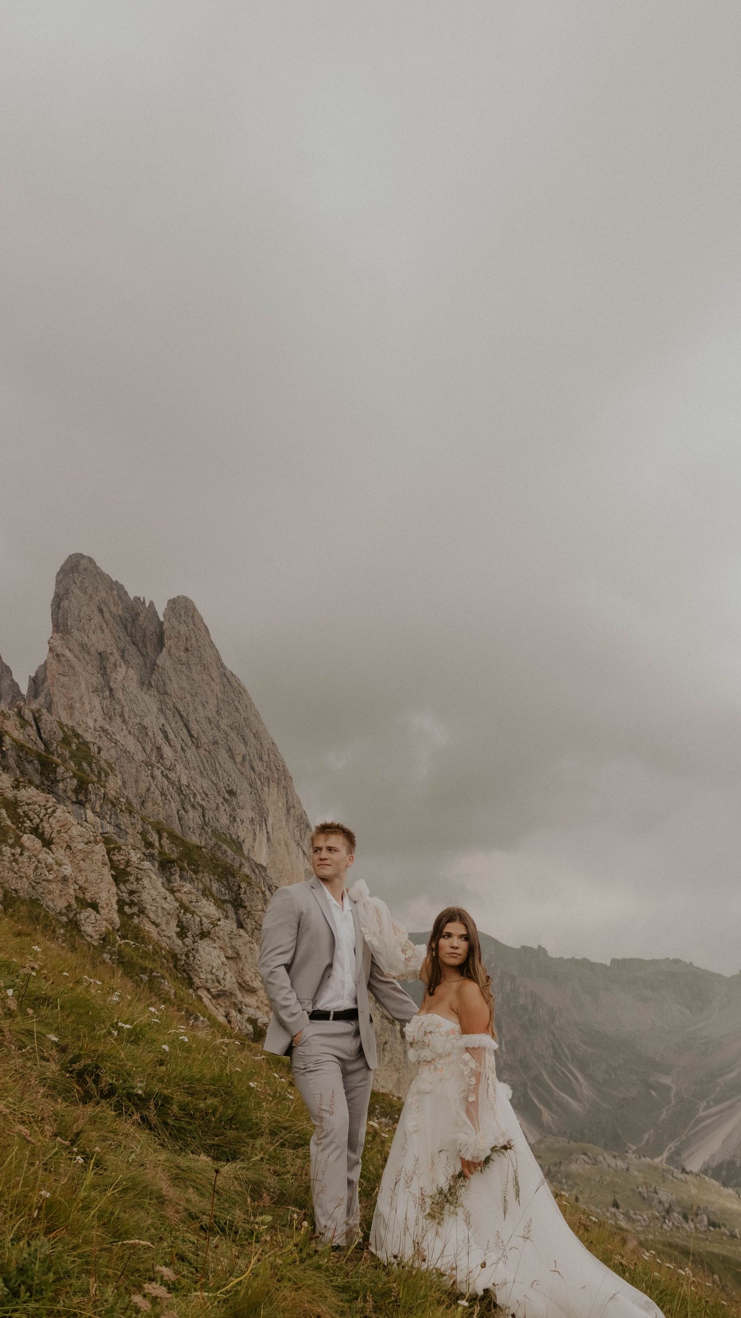 A couple in wedding attire poses on a mountainside with dramatic rocky peaks and cloudy skies in the background.