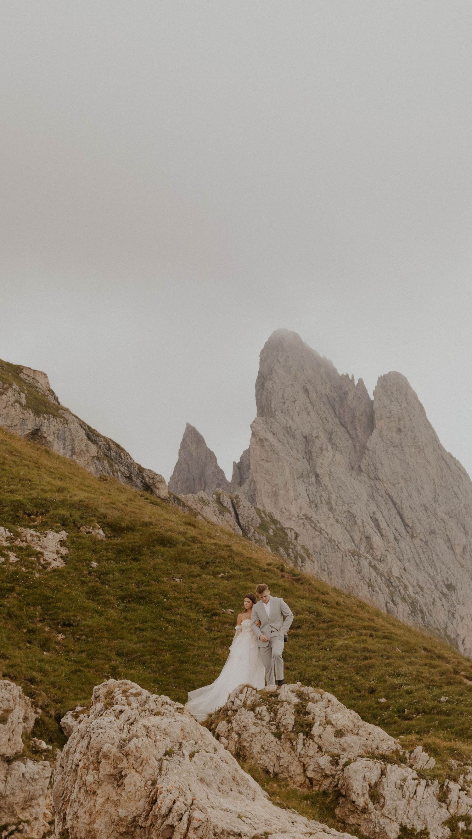 A couple stands on a rocky mountain peak in the Dolomites with dramatic jagged peaks in the background.