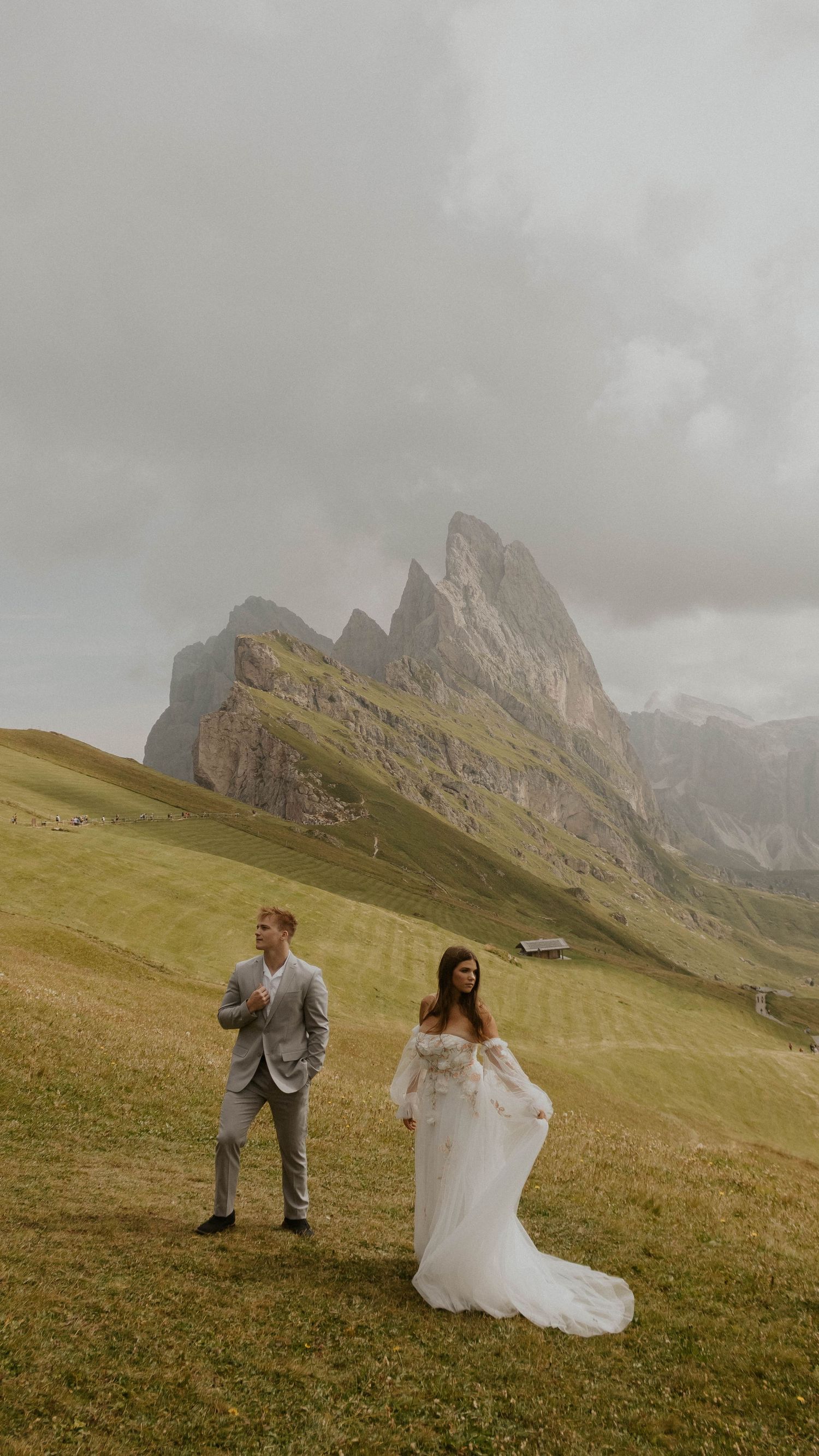 A couple in wedding attire poses on a grassy hillside with dramatic Dolomite mountain peaks rising in the misty background.