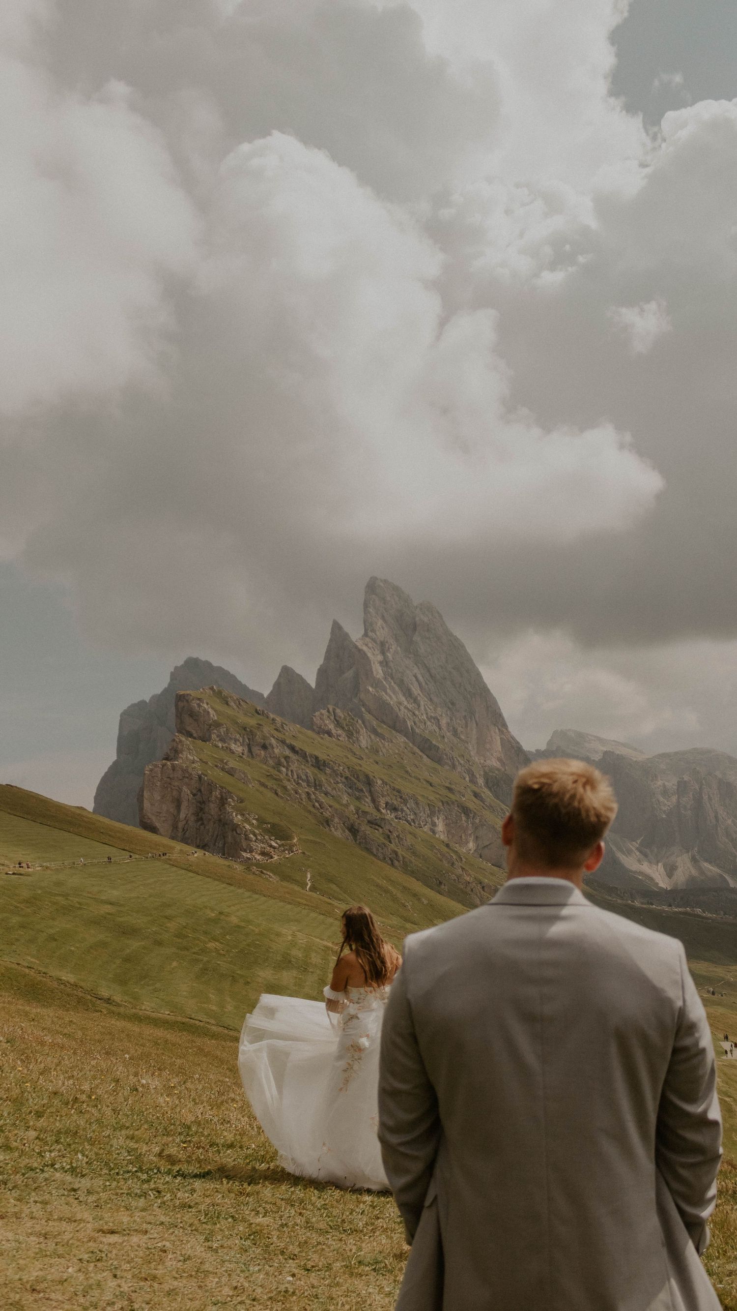 A couple in wedding attire walks across a grassy meadow with dramatic Dolomite mountain peaks rising in the background.