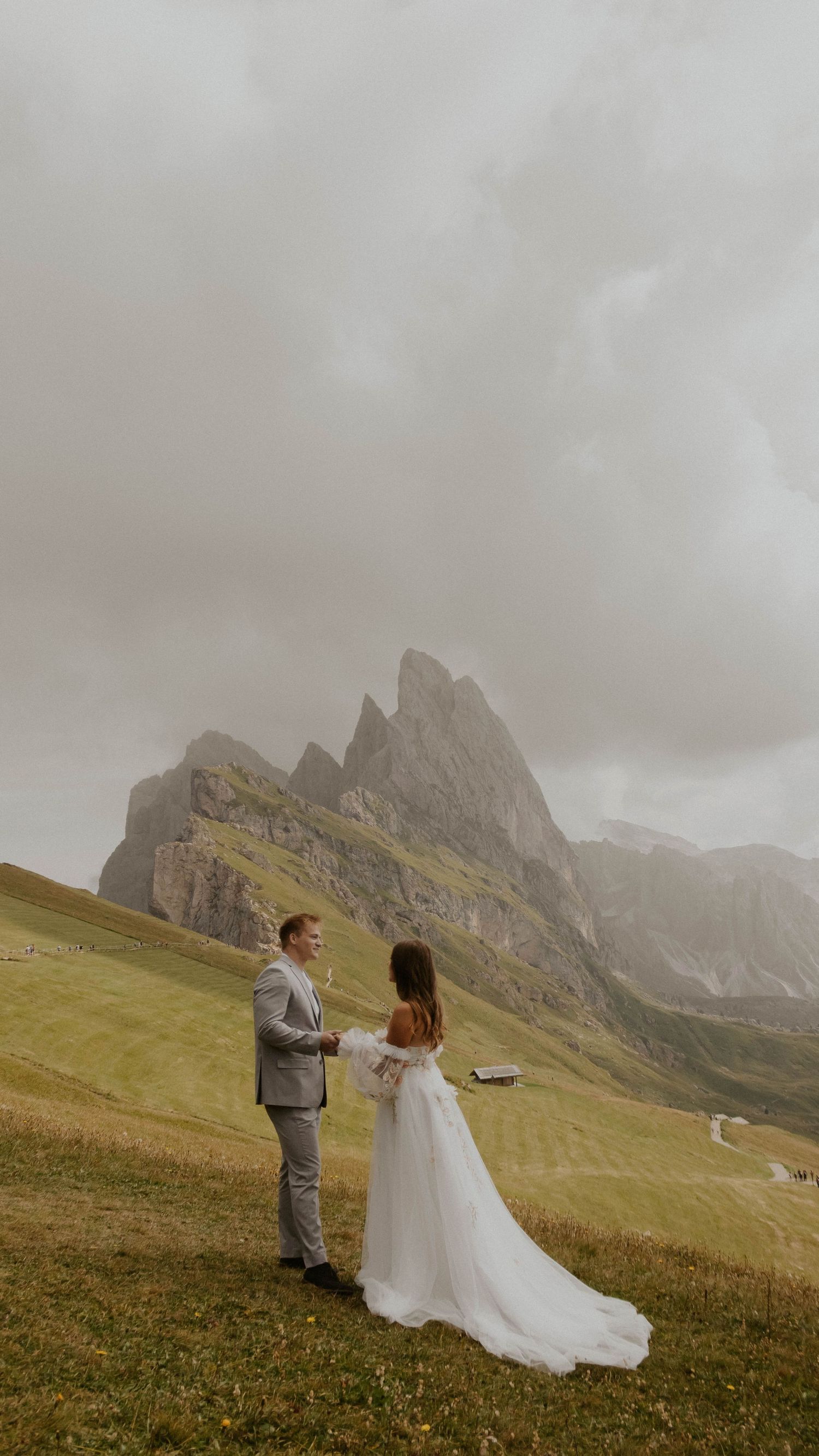 A couple in wedding attire stands on a grassy mountainside with dramatic Dolomite peaks rising in the misty background.