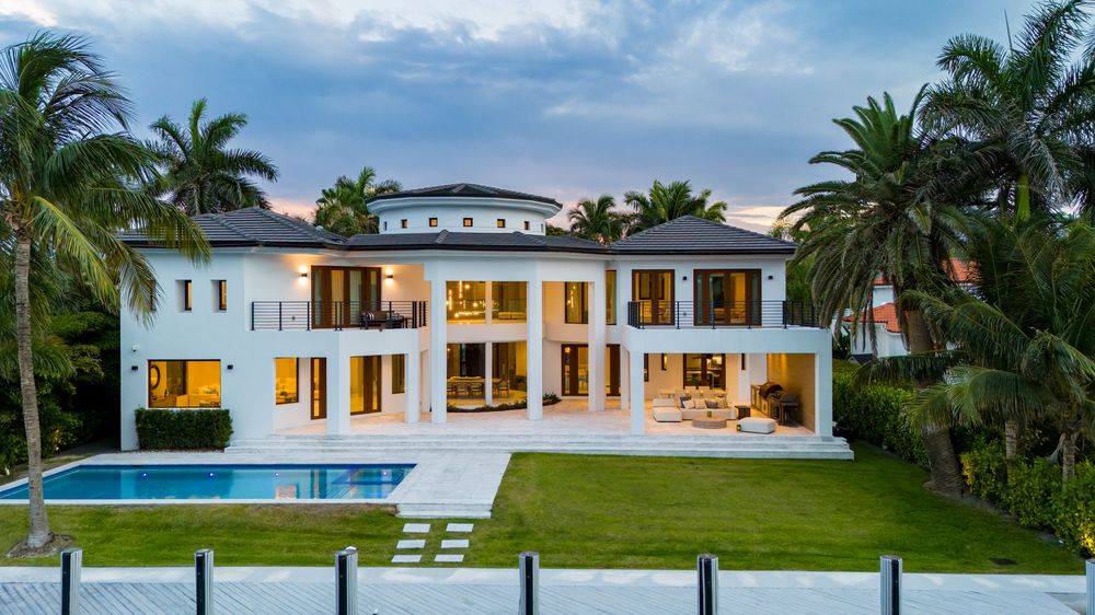 Panoramic twilight view of a modern luxury mansion with illuminated windows and palm trees in the tropical landscape.