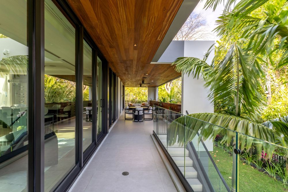 Long covered walkway with wooden ceiling and glass walls overlooking lush tropical gardens.