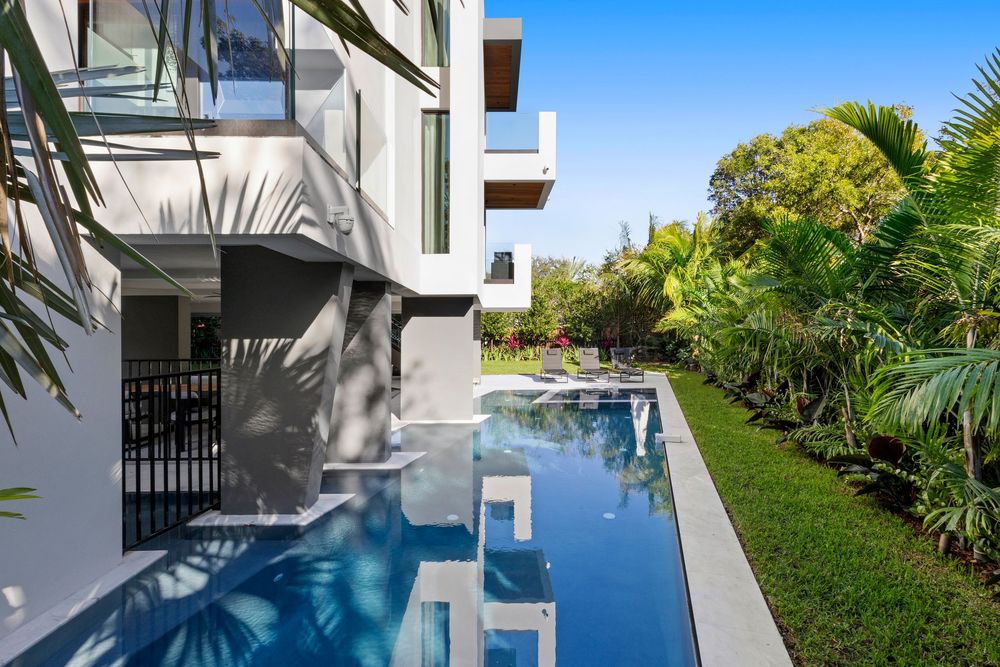 Infinity pool with clear blue water extending along the modern white house featuring tropical landscaping.