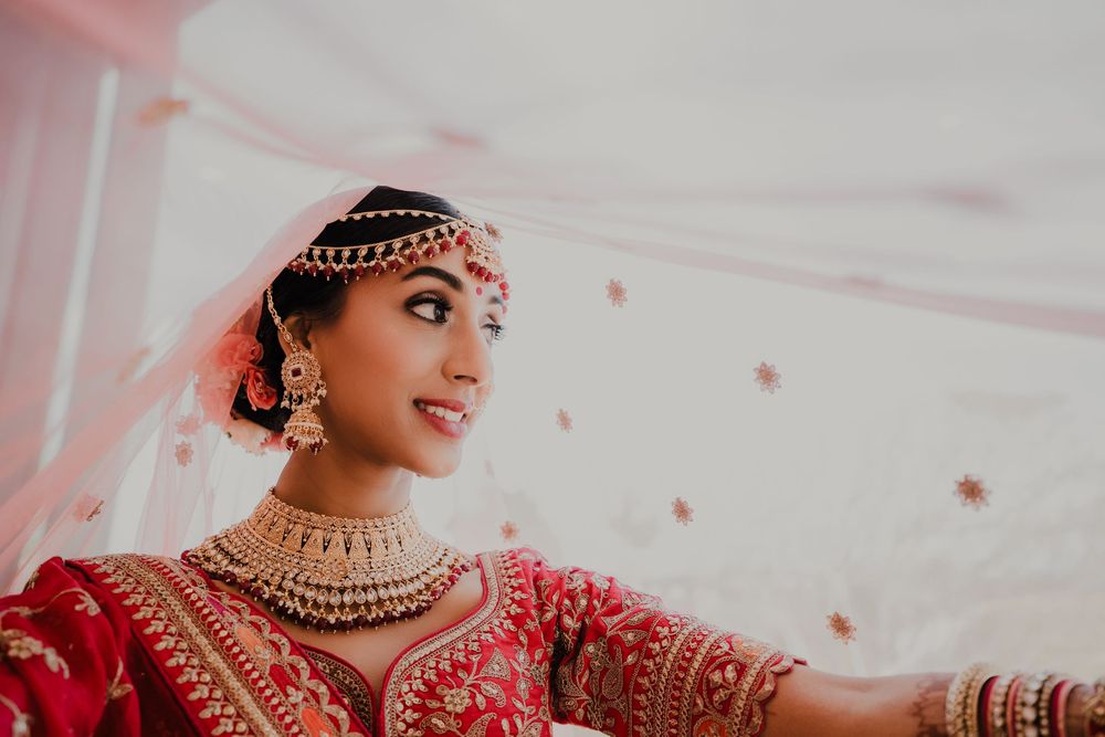 Close-up details of ornate gold and red Indian bridal jewelry and attire against a soft pink background.