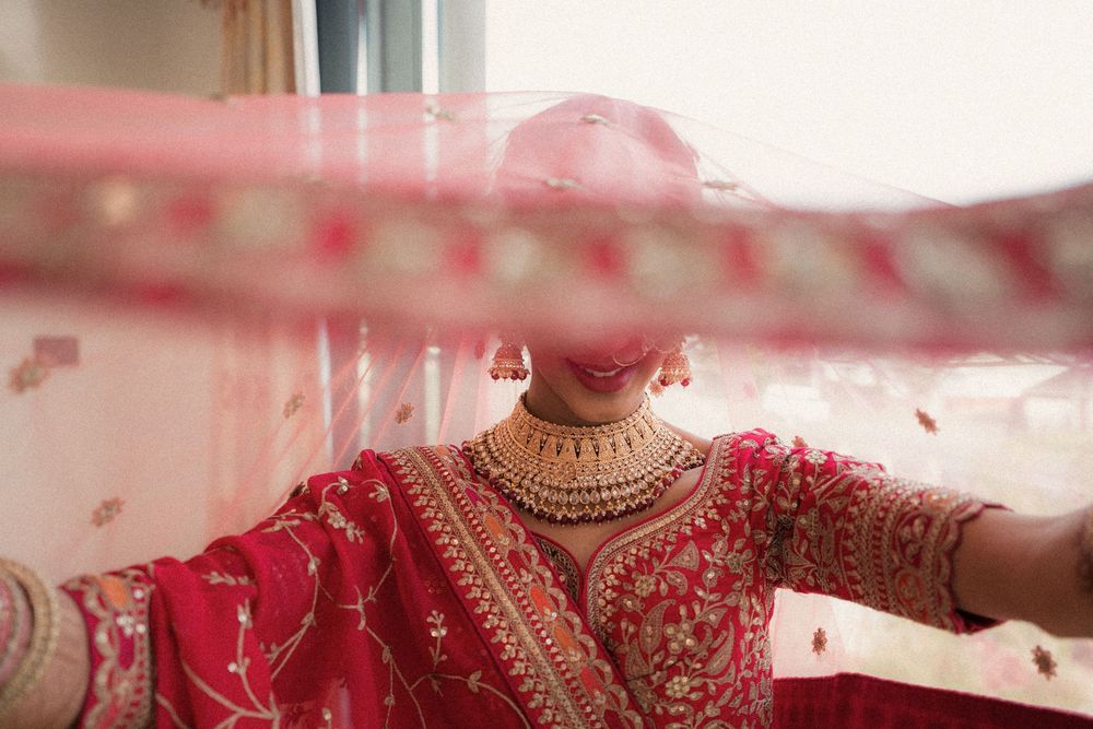 Traditional Indian bride in red embroidered sari with gold jewelry seen through flowing veil during wedding ceremony.