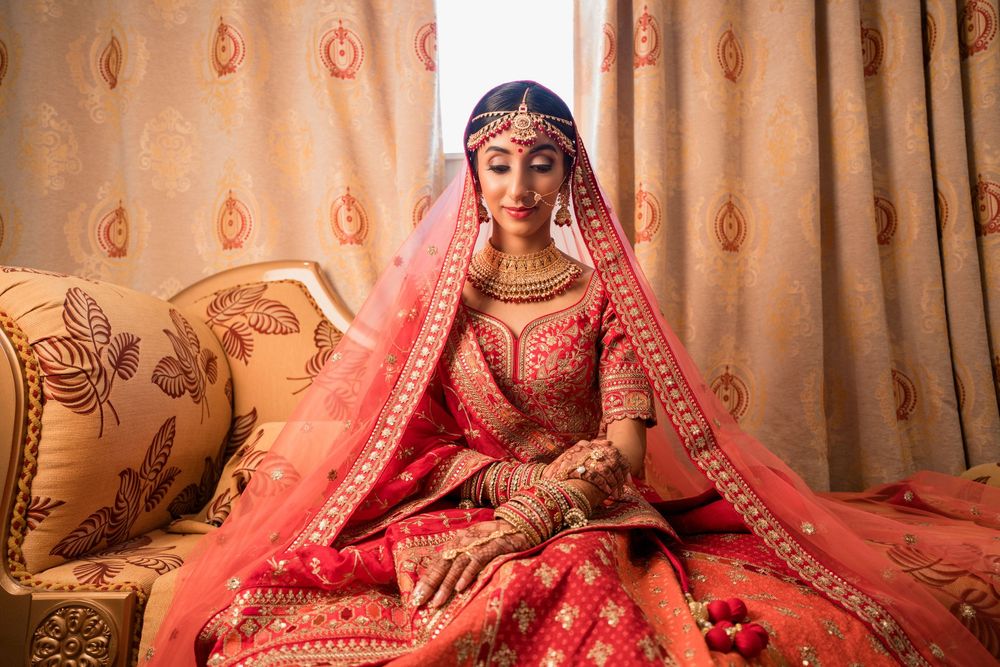 A person wearing a red bridal lehenga with intricate embroidery poses against a warm-toned backdrop.