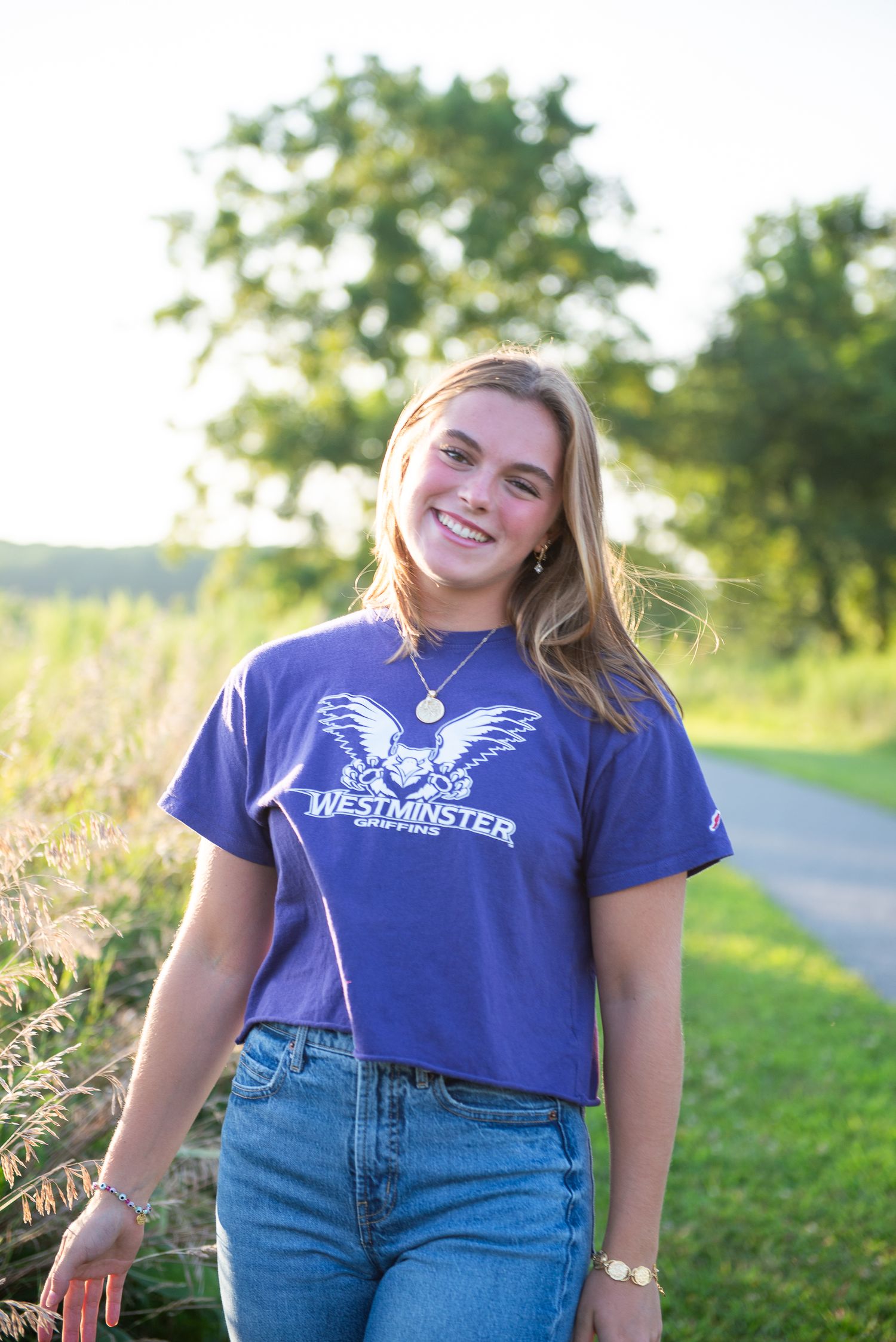 A person in a Westminster t-shirt and jeans stands outdoors on a sunny path next to tall grass and trees.
