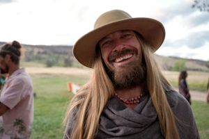 Person wearing a wide-brimmed hat smiles outdoors with mountains in background.