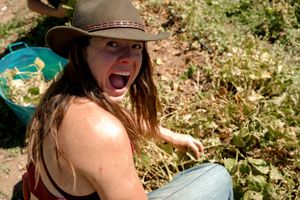 A person in a brown hat smiles enthusiastically while working outdoors in a garden.