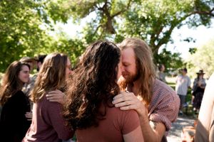 People share emotional embraces during an outdoor gathering under green trees.