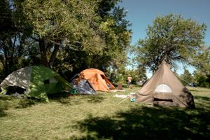 Several camping tents including an orange tent and teepee-style tent set up on grassy terrain under trees.
