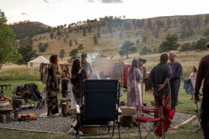 Outdoor gathering with people around camping chairs and grill with smoke rising in mountain setting.