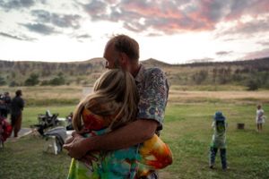 People embracing in a field at sunset with mountains and colorful sky in the background.