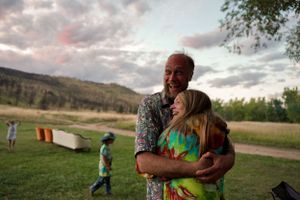 People sharing a warm embrace at sunset in a grassy field with mountains in the background.